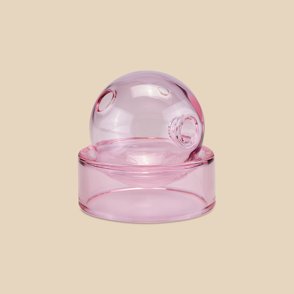 Seitlicher Blick auf die kugelrunde Crystal Ball Pipe in Pink, die auf einem farblich passenden Glasfuß steht. (Bild: AURIEY)